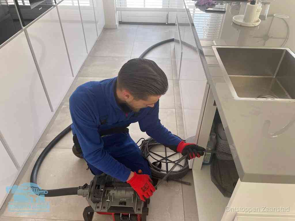 Een monteur ontstopt de keukenafvoer onder de spoelbak met een elektrische veermachine in Zaanstad