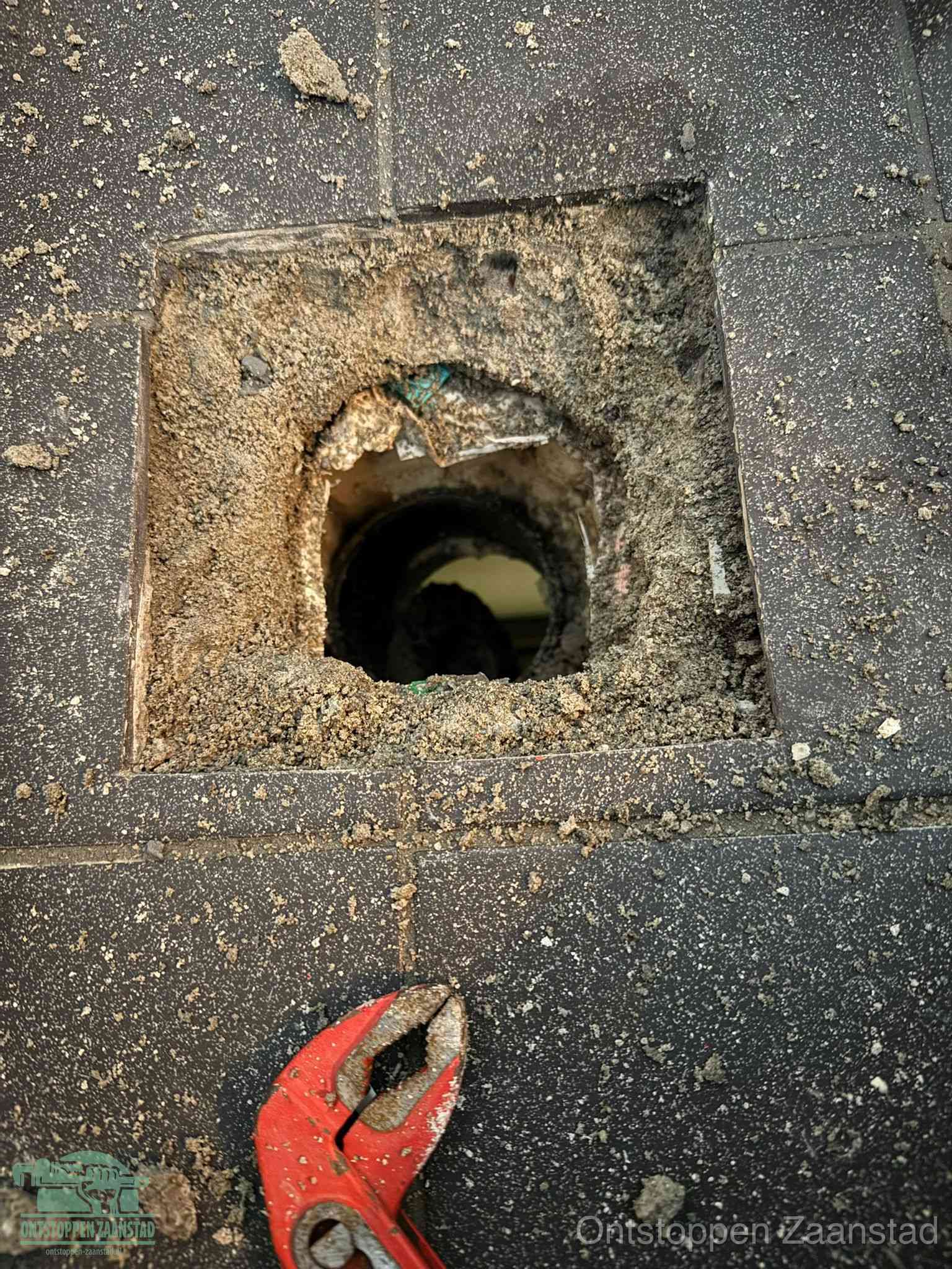 Een geopend rioolputje in een betontegelvloer met losgemaakt rooster en een zichtbare ronde afvoerbuis, het achterhalen waar de afvoerverstopping zit in Zaanstad