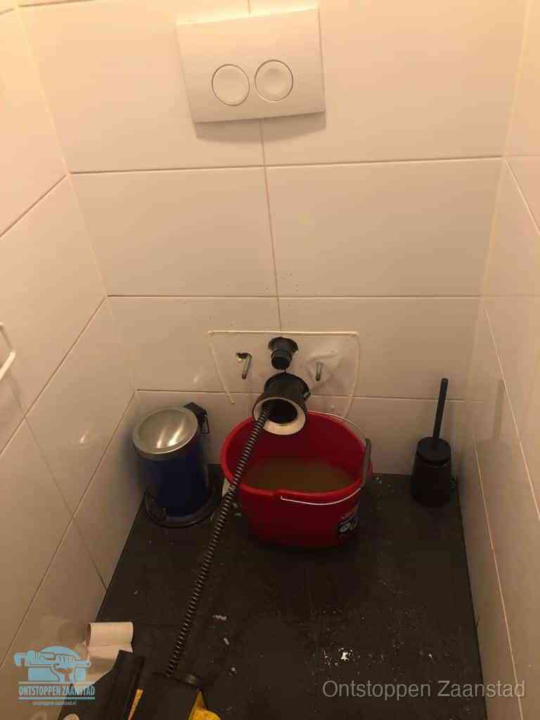 Een ontstopte wand-wc met verwijderde toiletpot waar een ontstoppingsveer in de wanduitgang verdwijnt, het achterhalen waar de verstopping zit in Zaanstad