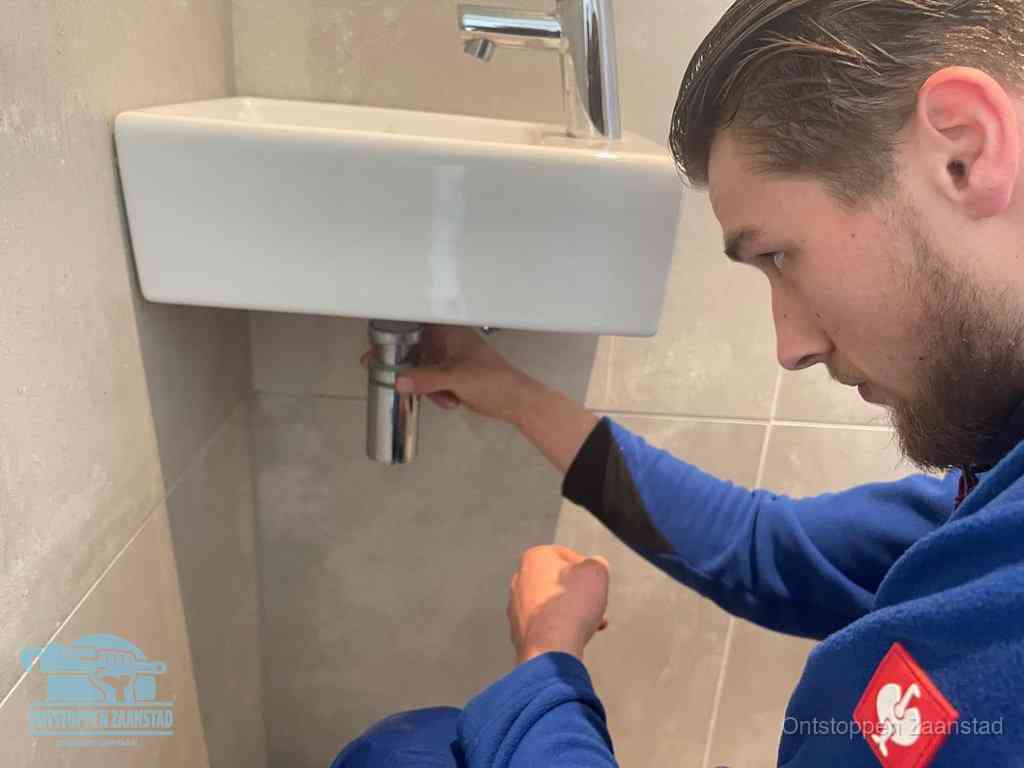 Een monteur draait met de hand de zwanenhalssifon onder een fonteinwasbak in de toiletruimte los om de bron van rioollucht in Zaanstad te vinden