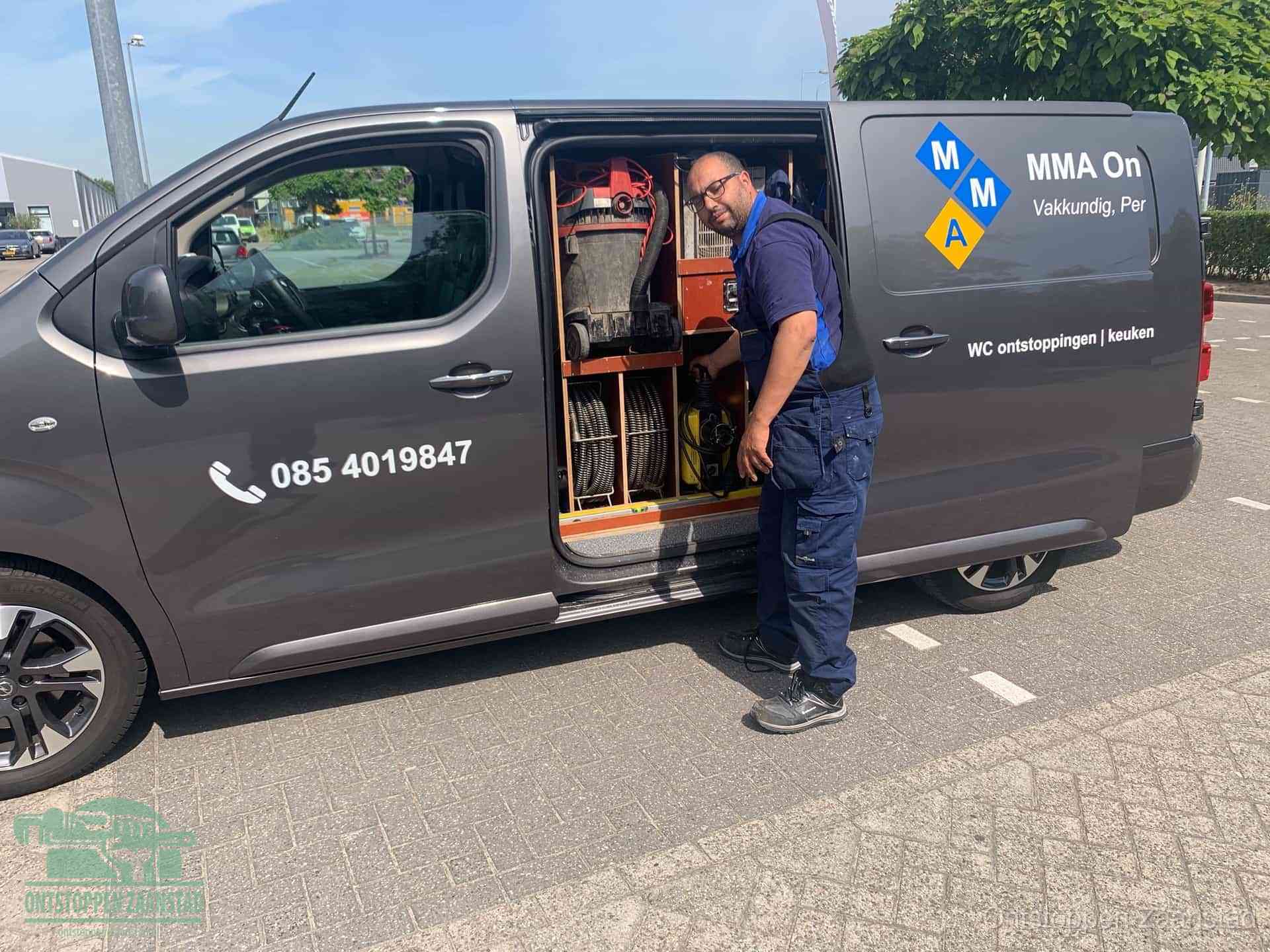 Een monteur bij de open schuifdeur van zijn bedrijfsbus op een parkeerplaats, klaar om de oorzaak van terugkerende afvoerverstoppingen aan te pakken in Zaanstad