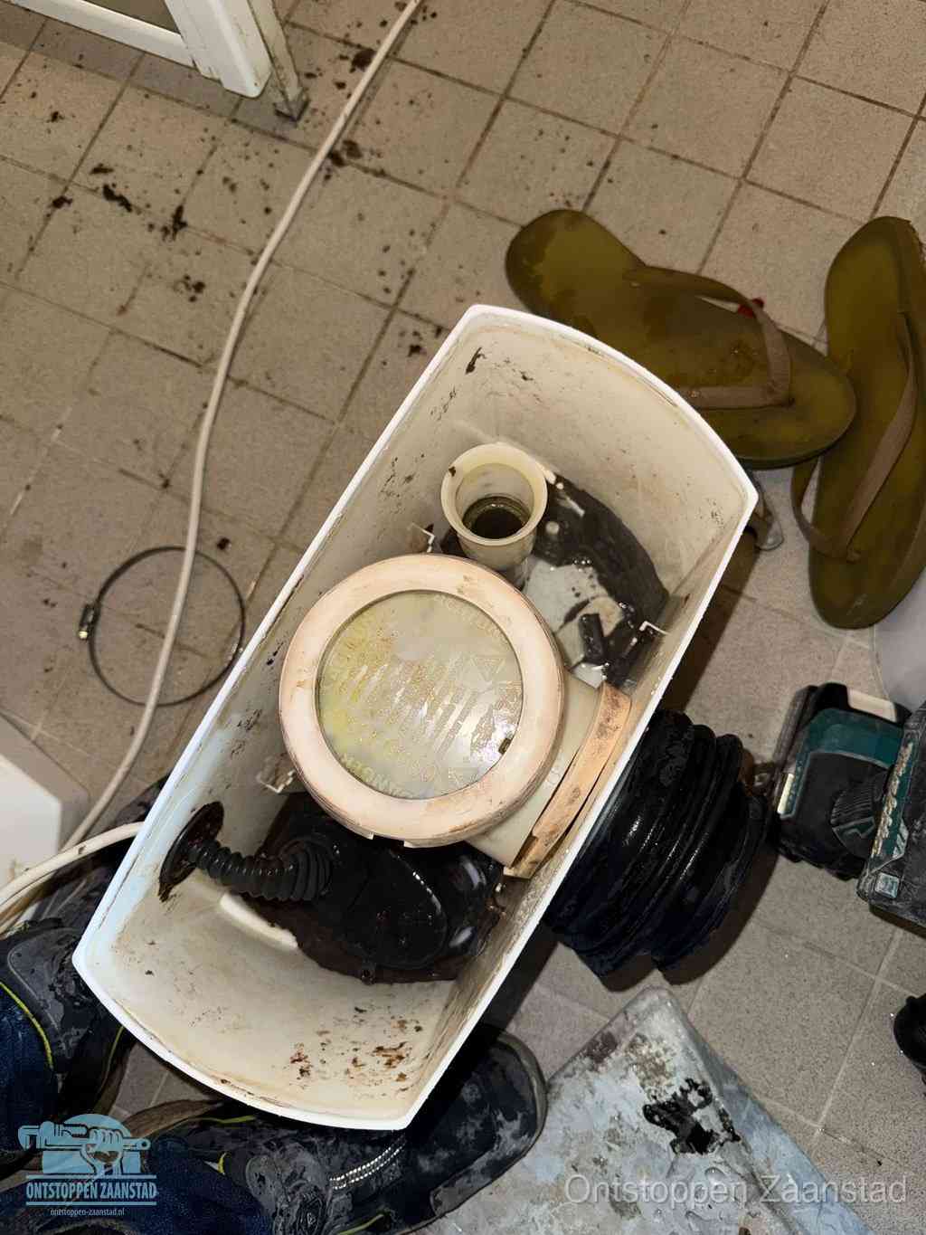Een gedemonteerde toiletpot op de vloer met ontstoppingsveer en gereedschap ernaast, het verwijderen van een vastzittend voorwerp uit de wc in Zaanstad