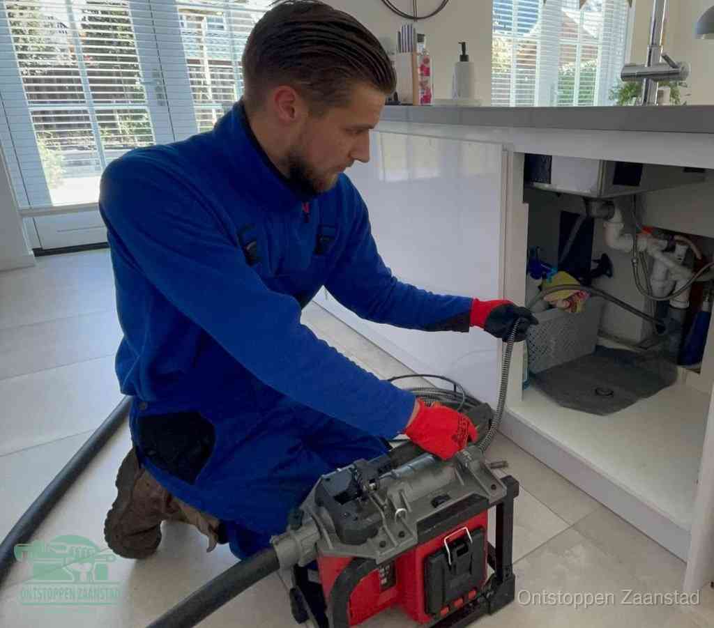 Een monteur met rode handschoenen knielt in een keuken en voert een elektrische ontstoppingsveer in tijdens het professioneel afvoer ontstoppen in Zaanstad