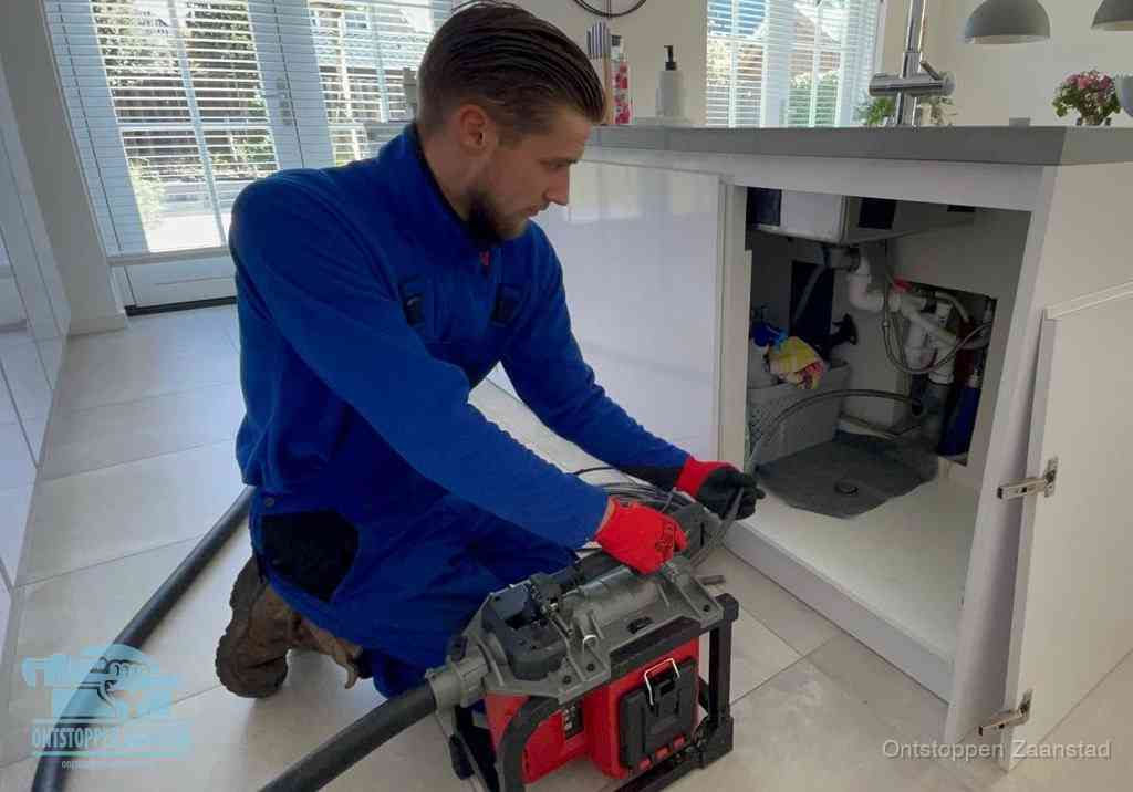 Een monteur voert een professionele elektrische ontstoppingsmachine in de afvoer onder de spoelbak tijdens het gootsteen ontstoppen in Zaanstad